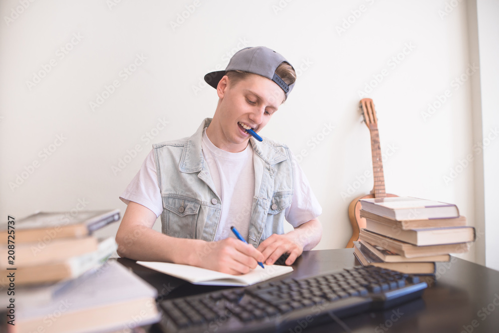Portrait of a smiling teenager sitting at the table, writing homework in a notebook and smiling. Student sitting at a work desk near books at home and writing in a notebook Portrait of a smiling teenager sitting at the table, writing homework in a notebook and smiling. Student sitting at a work desk near books at home and writing in a notebook