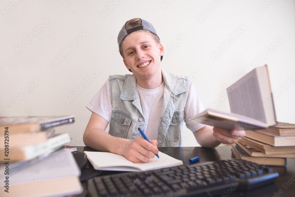 Portrait of a happy student sitting at the table with a book in his hands looking at the camera and smiles. A teenager with a pen and book in his hand makes a homework at home in the room Portrait of a happy student sitting at the table with a book in his hands looking at the camera and smiles. A teenager with a pen and book in his hand makes a homework at home in the room