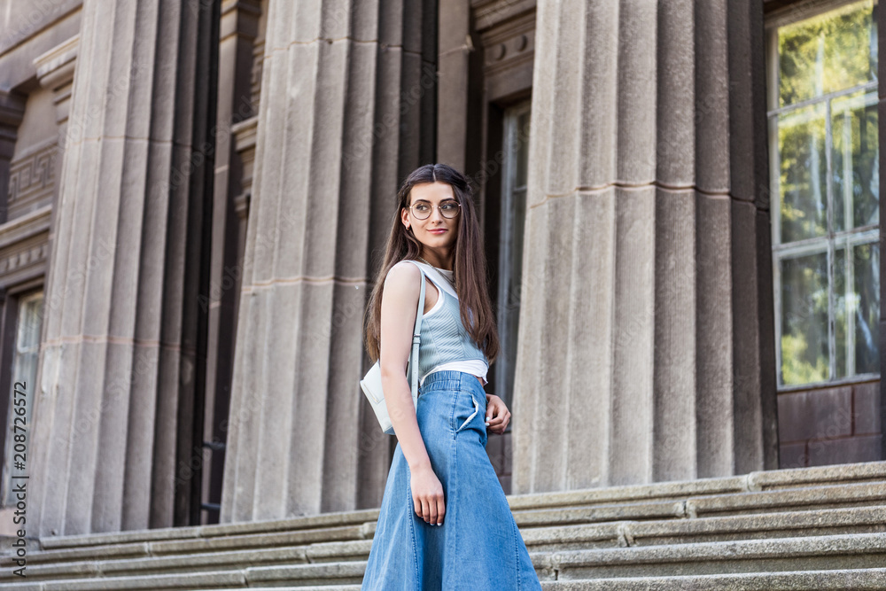 young stylish woman in eyeglasses and denim skirt standing on steps
