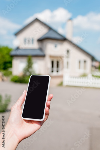 cropped shot of young woman holding smartphone with blank screen in front of house