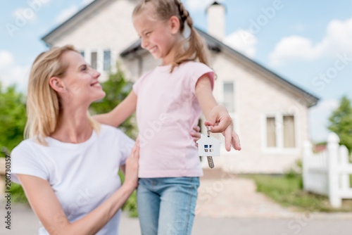closeup shot of key with trinket in hand of smiling little child embracing mother in front of new house