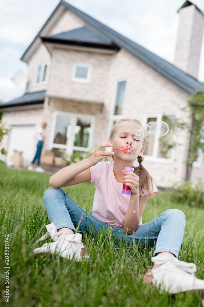 little kid using bubble blower and sitting on lawn while her mother standing behind in front of house