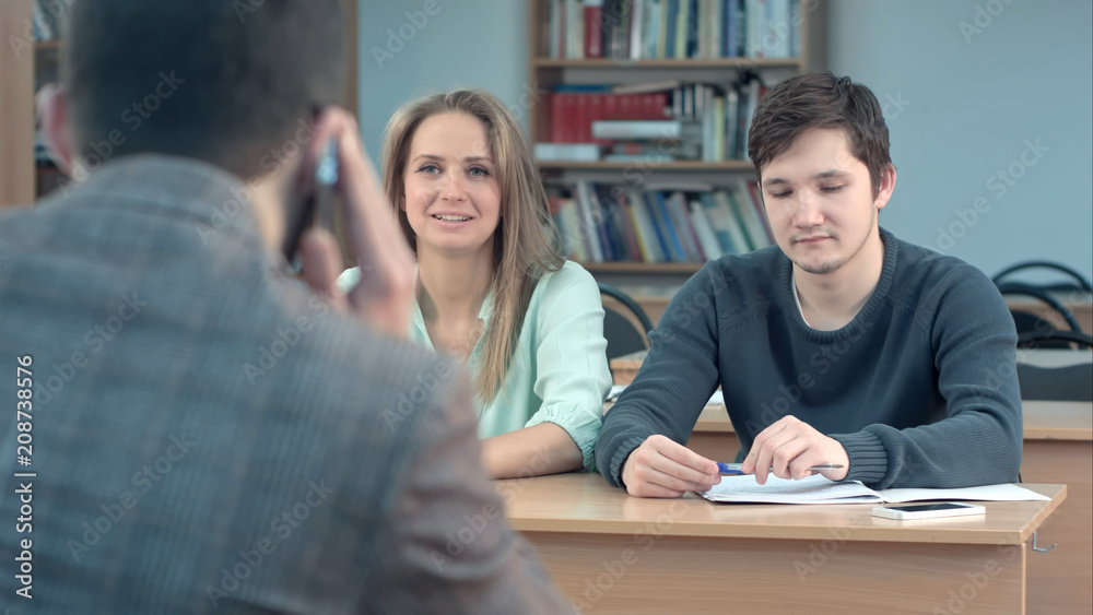 Group of university students sitting at their desks in auditorium and waiting for teacher, while he talking on smartphone Group of university students sitting at their desks in auditorium and waiting for teacher, while he talking on smartphone