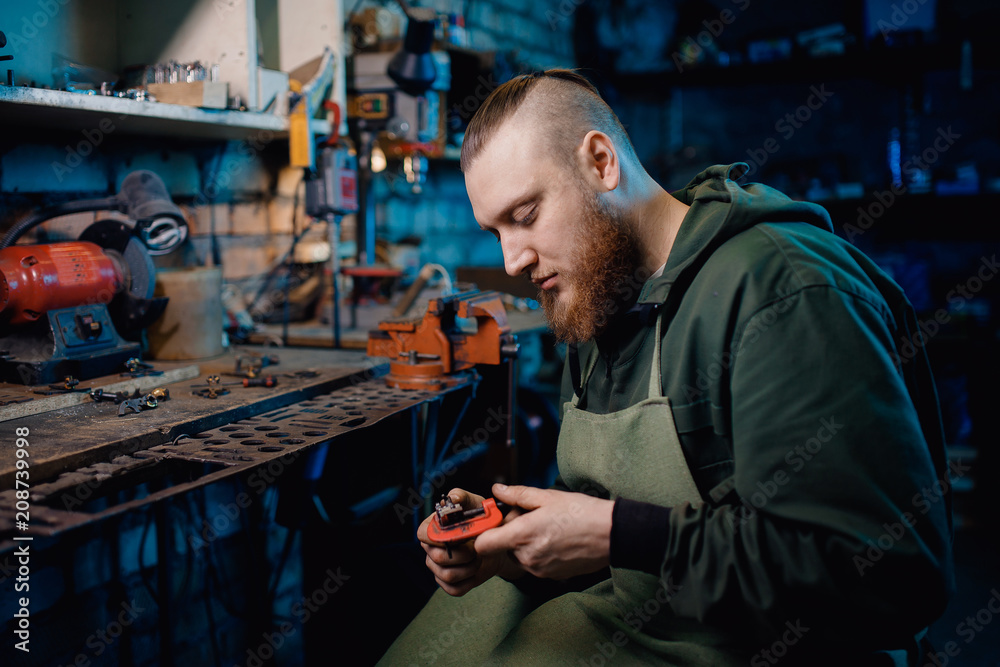 Man with beard in overalls makes details of iron and wood in garage workshop, handmade production. concept is professional jeweler. Man with beard in overalls makes details of iron and wood in garage workshop, handmade production. concept is professional jeweler.