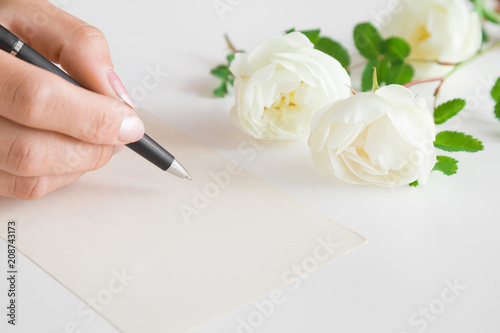 Woman s hand with pen writing on the white blank greeting card on the table. White roses. Fresh flowers.