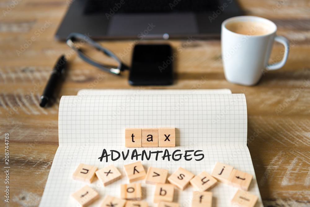 Closeup on notebook over vintage desk background, front focus on wooden blocks with letters making Tax Advantages text