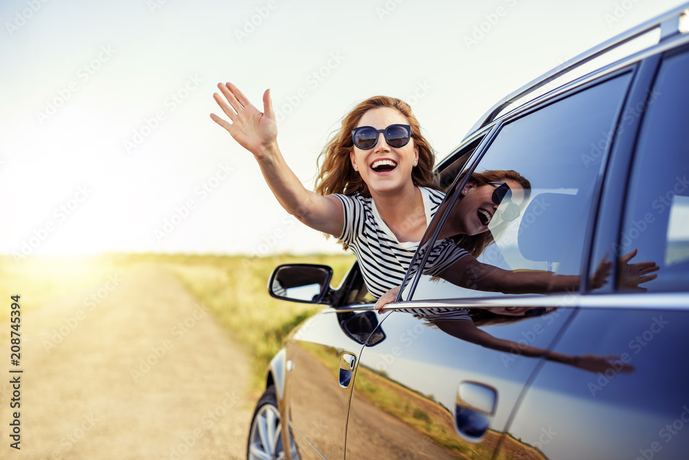 Attractive smiling woman waving her hand from the car window on a summer day.