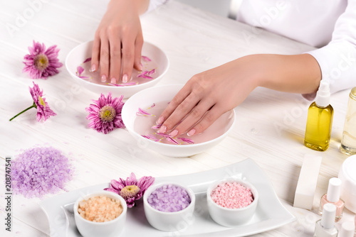 partial view of woman receiving bath for nails at table with flowers, colorful sea salt, cream container, aroma oil bottles and nail polishes in beauty salon