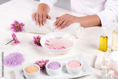 cropped image of woman holding hands on towel for manicure procedure at table with flowers, colorful sea salt, cream container, aroma oil bottles and nail polishes in beauty salon