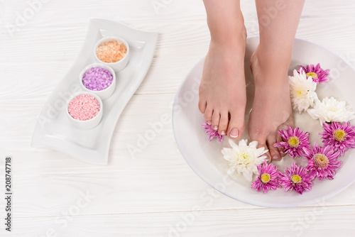 partial view of barefoot woman receiving bath for nails with sea salt and flowers in beauty salon