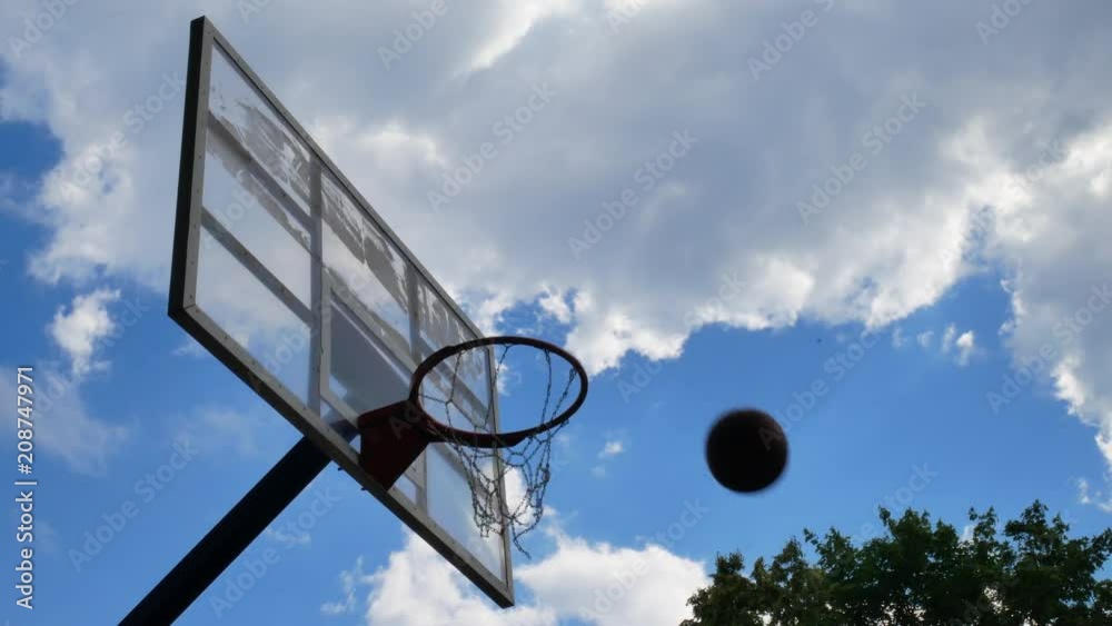 Basketball basket hoop with ball going through net at street basketball sport arena at blue sky with white clouds background Basketball basket hoop with ball going through net at street basketball sport arena at blue sky with white clouds background