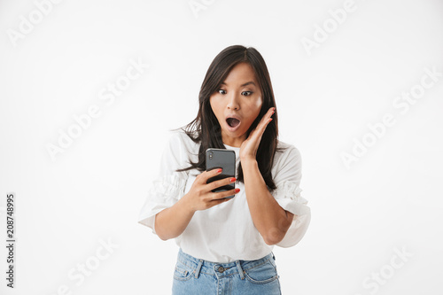 Image of shocked or surprised asian woman screaming and gesturing in panic, while looking at smartphone, isolated over white background