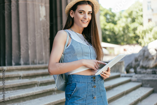 beautiful young woman in hat using digital tablet and smiling at camera while standing on stairs
