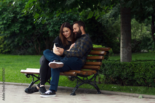 Happy young couple sitting in a park bench and using a smart phone