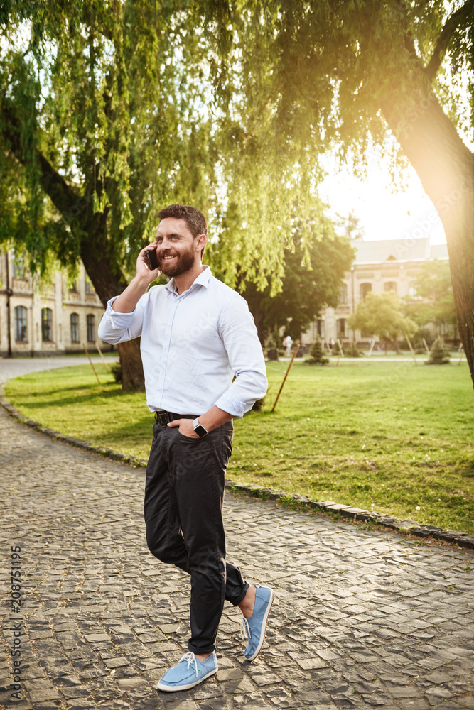 Full length photo of smiling stylish man 30s, walking through park on sunny day and having mobile call Full length photo of smiling stylish man 30s, walking through park on sunny day and having mobile call