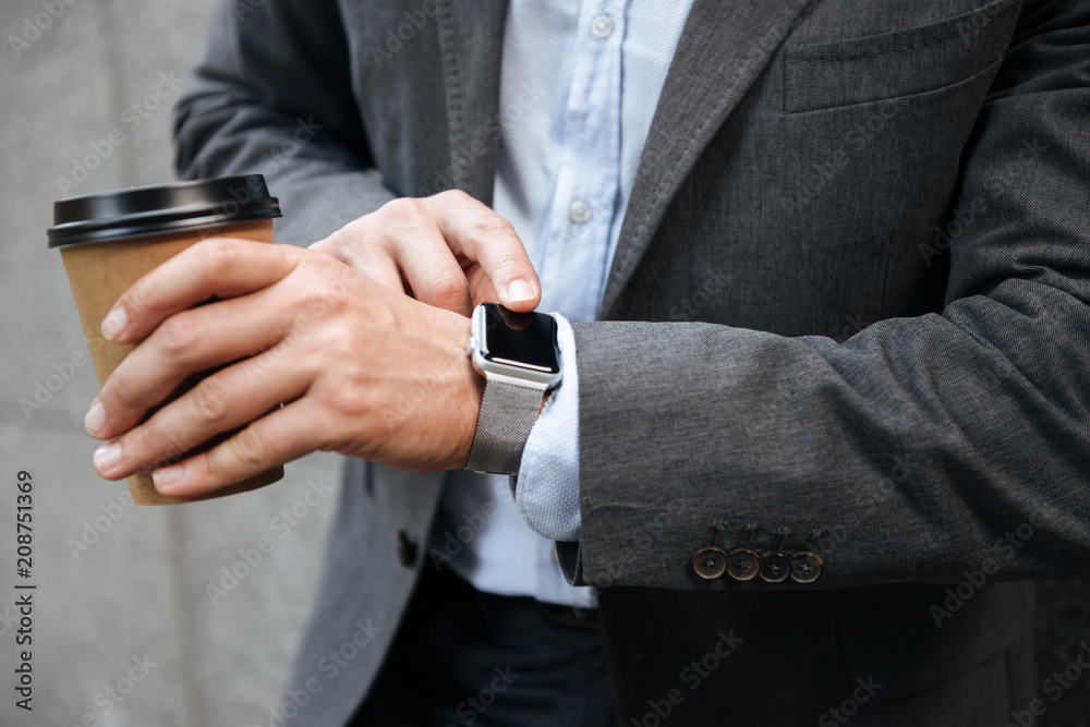 Cropped photo of european businessman in gray suit and white shirt standing against granite wall, and touching smartwatch while drinking takeaway coffee Cropped photo of european businessman in gray suit and white shirt standing against granite wall, and touching smartwatch while drinking takeaway coffee