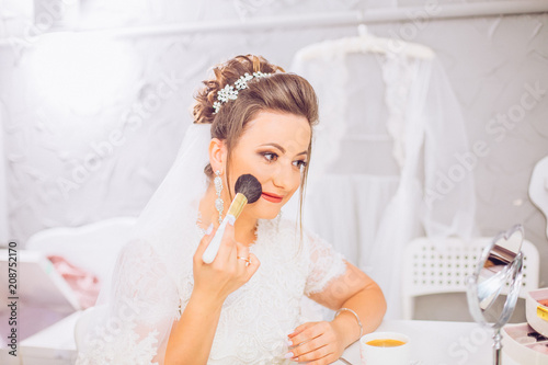 Attractive young bride preparing for wedding in the morning, drinking coffee with smatphone smiling and looking to the mirror. Bride waiting for the groom. Wedding concept.