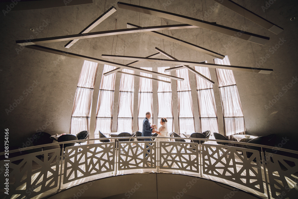 Obraz premium Newlyweds stand against the background of large windows and are reflected in them, in the background of a light bulb and a bokeh. The bride is in a lace dress. Stylish wedding in the interior.