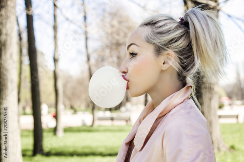 Obraz na plátně Young woman blowing bubble with chewing gum and walking in park outdoors, lifest