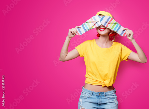 Young redhead girl in yellow t-shirt and blue jeans holding a summer flip flops sandals on pink background.