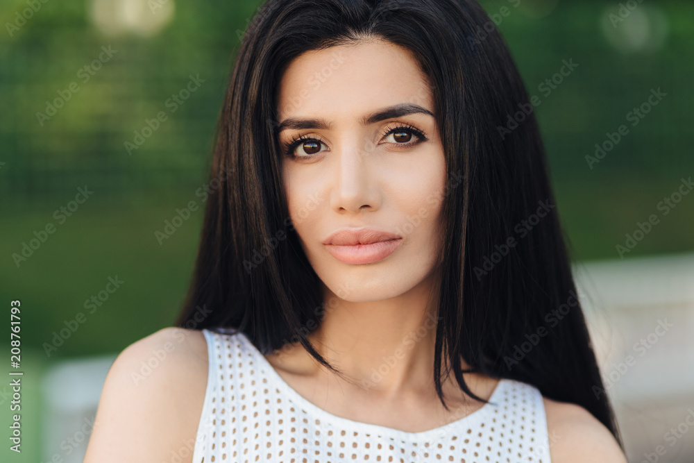 Gorgeous young woman with straight dark hair, has appealing appearance, looks confidently at camera, poses for fashion magazine, breathes fresh air outdoor, poses against green nature background Gorgeous young woman with straight dark hair, has appealing appearance, looks confidently at camera, poses for fashion magazine, breathes fresh air outdoor, poses against green nature background