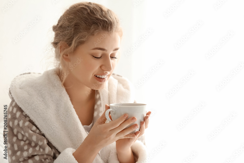 Young woman in robe drinking tea on light background