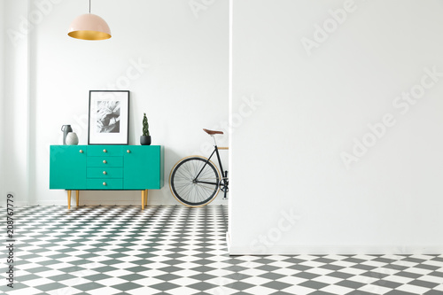 Empty wall and turquoise cabinet with decorations standing next to a bike in a hallway interior with checkered floor. Place for your poster or furniture