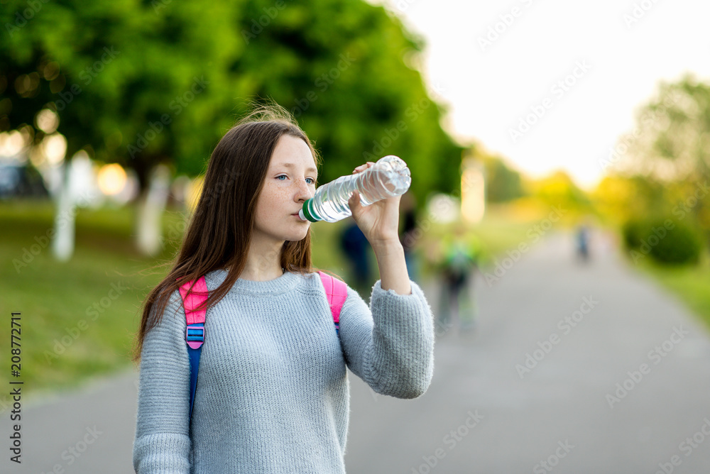 Beautiful schoolgirl girl. Summer nature. He holds a bottle of water and drinks. Brunette with long hair. Behind the back is a school backpack with textbooks. Green street in the city. Beautiful schoolgirl girl. Summer nature. He holds a bottle of water and drinks. Brunette with long hair. Behind the back is a school backpack with textbooks. Green street in the city.