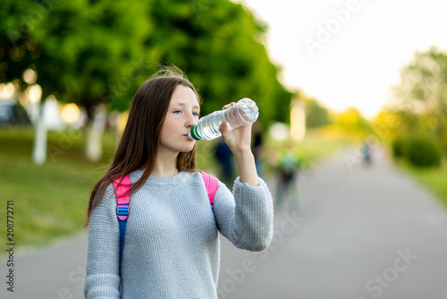 Beautiful schoolgirl girl. Summer nature. He holds a bottle of water and drinks. Brunette with long hair. Behind the back is a school backpack with textbooks. Green street in the city.