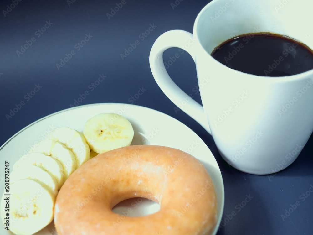 close up from black coffee in white coffee cup with soft focus (banana and donut) at foreground put on blue background close up from black coffee in white coffee cup with soft focus (banana and donut) at foreground put on blue background