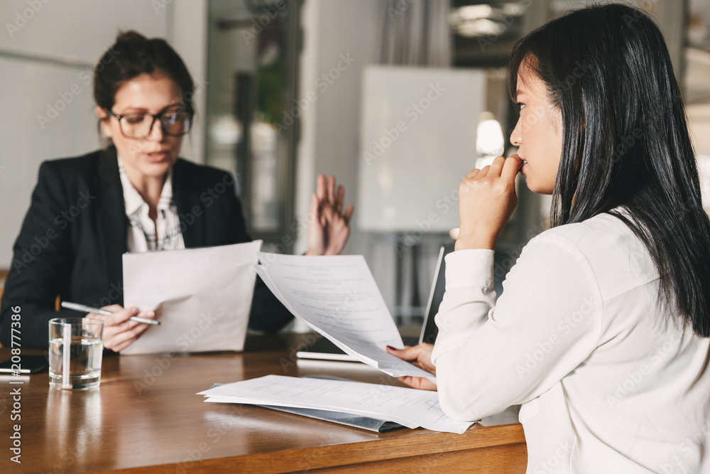 Image of nervous asian woman looking and talking to businesswoman, while sitting at table in office during job interview - business, career and recruitment concept Image of nervous asian woman looking and talking to businesswoman, while sitting at table in office during job interview - business, career and recruitment concept