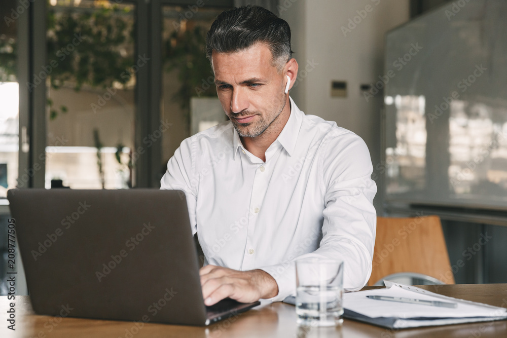 Photo of european businessman 30s wearing white shirt and wireless earphones sitting at table in office, and working at laptop