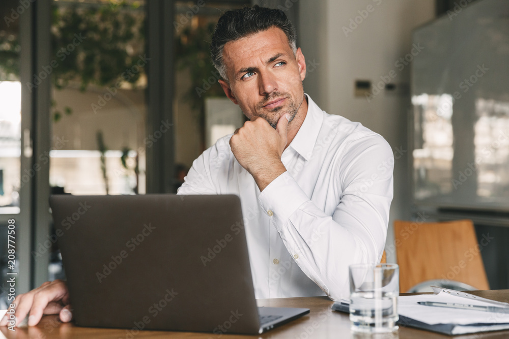 Photo of european man wearing white shirt and earbud sitting at table in office with brooding look aside, while working on laptop