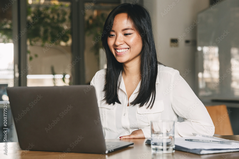 © Drobot Dean - Image of joyful asian female worker 20s wearing white shirt smiling while sitting at table in office, and working on laptop