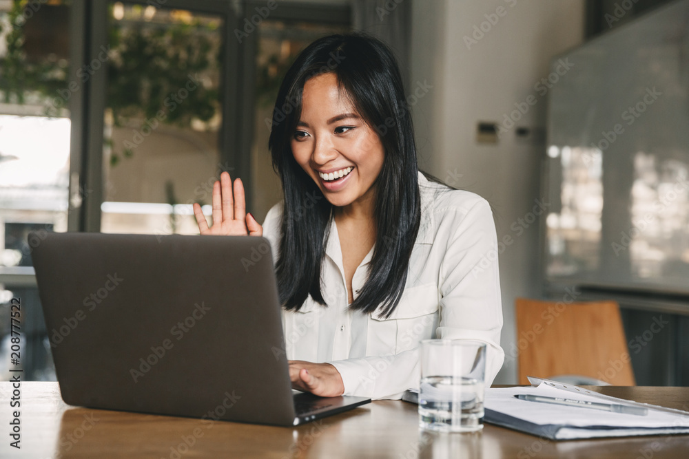 Image of happy asian woman 20s wearing white shirt smiling and waving hand at laptop, while speaking or chatting on video call in office Image of happy asian woman 20s wearing white shirt smiling and waving hand at laptop, while speaking or chatting on video call in office