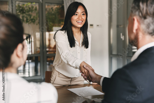 Business, career and placement concept - image from back of two employers sitting in office and shaking hand of young asian woman, after successful negotiations or interview photo