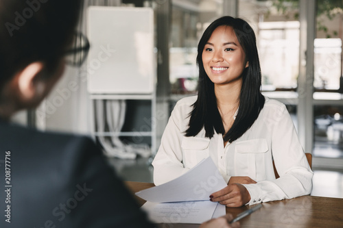 Portrait of joyful asian woman smiling and holding resume, while sitting in front of businesswoman during corporate meeting or job interview - business, career and placement concept