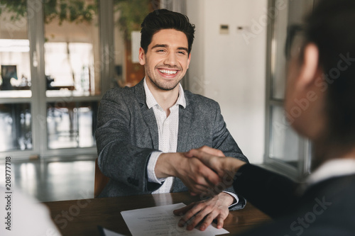 Business, career and placement concept - happy caucasian man 30s rejoicing and shaking hands with employee, when was recruited during interview in office photo