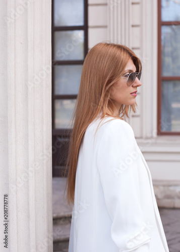 A woman in a white jacket and sunglasses looks away from the column. Street style. Fashion