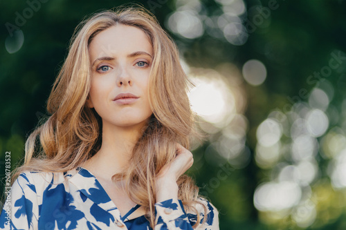 Horizontal shot of pretty blue eyed gorgeous female with long wavy hair, has thoughtful expression, poses against green blurred background with copy space for your advertisement. People and leisure