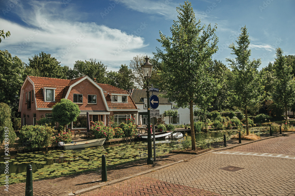 Tree-lined canal with aquatic plants, boats on the banks and brick houses on a sunny day in Weesp. Quiet and pleasant village full of canals and green near Amsterdam. Northern Netherlands.