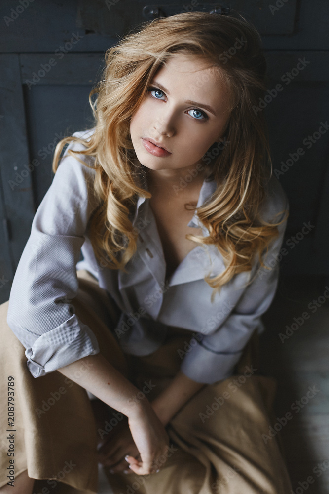 Fototapeta premium Fashionable and beautiful blonde model girl with light makeup and amazing blue eyes in unbuttoned blue shirt and in khaki pants sits and posing in art interior