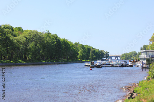 City Park Scene with Yacht Club Pier on Small Local River Water. Green Grass and Trees Summer Panoramic Outdoor Landscape View, Rural Park River with Boats on Sunny Day.