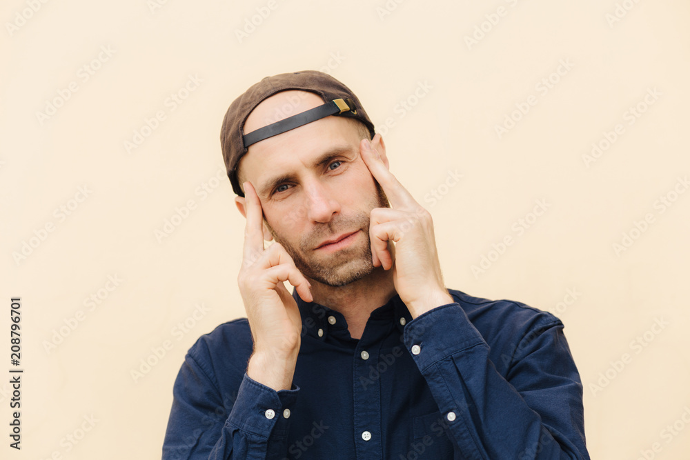 Candid shot of attractive unshaven male looks thoughtfully and concetrated at camera, keeps fore fingers on temples, wears stylish cap and shirt, isolated over light background. People and style Candid shot of attractive unshaven male looks thoughtfully and concetrated at camera, keeps fore fingers on temples, wears stylish cap and shirt, isolated over light background. People and style
