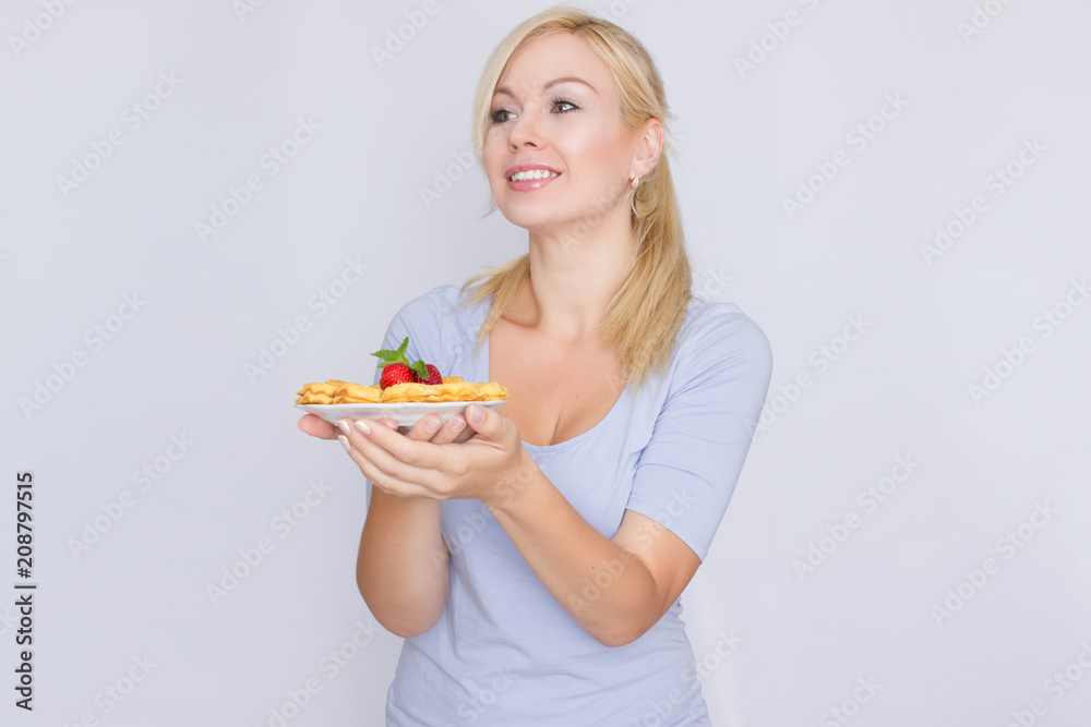 A happy blonde is holding a plate with Belgian waffles in her hands. Breakfast is decorated with strawberries and mint leaves. Concept: home food. A happy blonde is holding a plate with Belgian waffles in her hands. Breakfast is decorated with strawberries and mint leaves. Concept: home food.