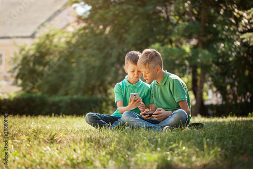 Wallpaper Mural Two kids brothers playing games on smartphone with excitement while sitting on grass in park. Torontodigital.ca