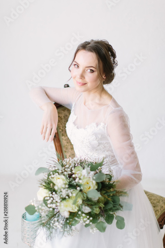 young beautiful bride sitting in a chair and smiling, close-up. Beauty portrait of bride wearing fashion wedding dress with luxury delight make-up and hairstyle, studio indoor photo.