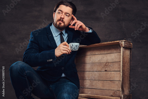 Pensive middle age businessman with stylish hair and beard dressed in jeans, jacket and tie, sitting on wooden boxes and holds a cup of coffee in a studio.
