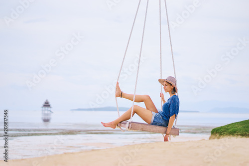 Vacation concept. Happy young woman in hat sitting on swing enjoying sea view.