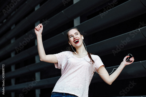 human emotion. portrait of a young cheerful girl on a gray background of the city building. the model listens to music on the phone with headphones  rejoices and dances.